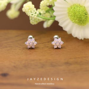 May include: A pair of flower-shaped stud earrings with a silver post and a cluster of light pink gemstones. The earrings are set on a wooden surface with floral accents in the background. The text "JAYZEDESIGN Handcrafted Jewellery" is at the bottom.
