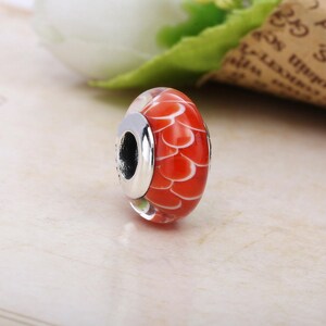 May include: A red and white glass bead with a silver-coloured metal core. The bead has a pattern of overlapping white shapes on a red background. The bead is likely for a bracelet or necklace.