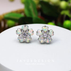 May include: A pair of flower-shaped stud earrings. Each earring features five opal petals surrounding a cluster of small, clear gemstones. The earrings are set in a silver metal. The text "JAYZEDESIGN Handcrafted Jewellery" is at the bottom.