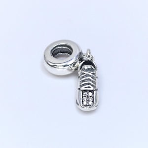 May include: A silver charm depicting a small trainer with detailed lace design and a sparkling toe. The charm is attached to a round, ring-like element, suitable for a bracelet or necklace. The charm is made of a shiny, metallic material.