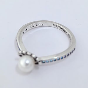 Pode incluir: Um anel de prata com uma pérola branca e detalhes azuis. O anel é marcado com "©Disney" e "PANDORA 925". O design é delicado, com pequenas pedras azuis ao longo da banda.
