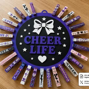 Peut inclure: Un présentoir mural circulaire violet et noir avec le texte CHEER LIFE en lettres violettes, un nœud blanc et un cœur blanc. Le pourtour est orné de nombreuses pinces à linge décorées avec divers accents violets, blancs et argentés.