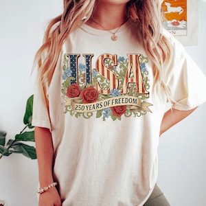 May include: Cream-colored t-shirt featuring the word "USA" in a vintage design with red and blue stripes and floral accents. Below the word "USA" is a banner that reads "250 YEARS OF FREEDOM".