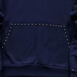 May include: Navy blue hoodie with a kangaroo pocket. The pocket and edges are embellished with small, silver-colored studs. The hoodie has a ribbed hem and cuffs, and the fabric appears to be a soft, medium-weight material.