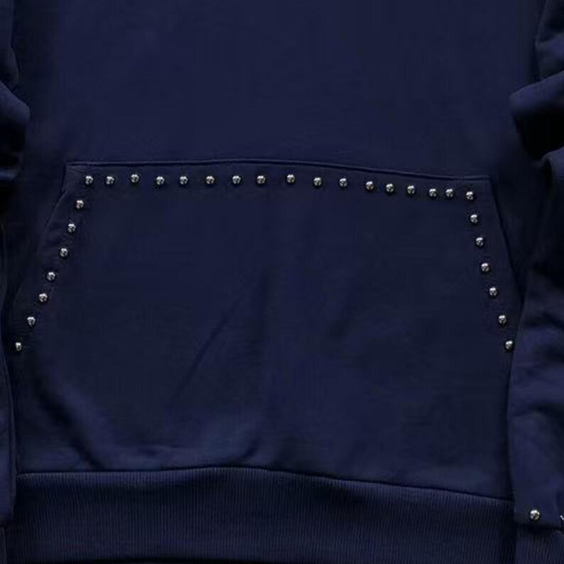 May include: Navy blue hoodie with a kangaroo pocket. The pocket and edges are embellished with small, silver-colored studs. The hoodie has a ribbed hem and cuffs, and the fabric appears to be a soft, medium-weight material.