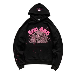 May include: Black hoodie with a pink spiderweb design and the text "666" in pink. The hood features a small pink crown graphic. Pink star accents are on the sleeves and lower front. A classic, comfortable sweatshirt.