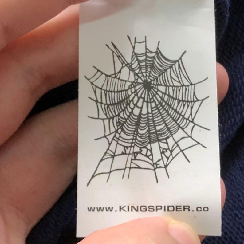 May include: A white tag with a black spiderweb design. The tag also includes the text "www.KINGSPIDER.co". The tag is rectangular and appears to be attached to a piece of clothing.