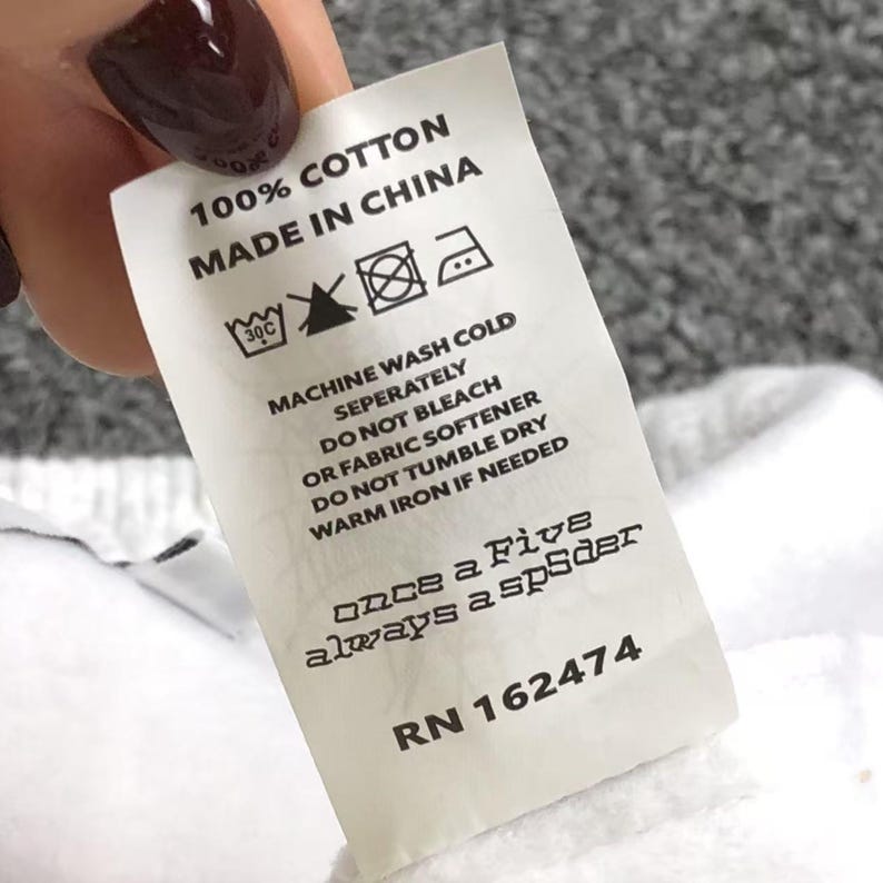 May include: A white clothing label with black text detailing fabric composition and care instructions. The label states "100% COTTON MADE IN CHINA". It also features the phrase "once a Five always a spider" and the RN number 162474.