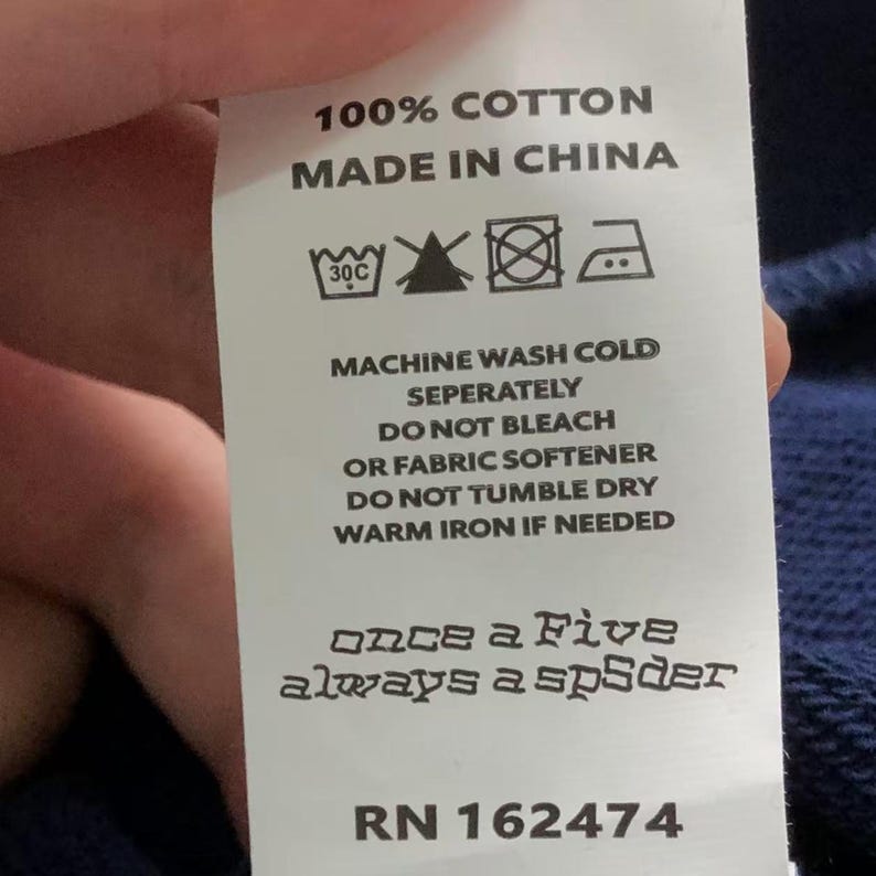 May include: A white clothing tag with black text. The tag reads "100% COTTON, MADE IN CHINA." Washing instructions include "MACHINE WASH COLD, DO NOT BLEACH." The tag also has the text "once a Five always a spSder" and the number "RN 162474."