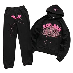 May include: Black sweatsuit set featuring a hoodie and sweatpants. The hoodie has a front pocket and a hood with a small graphic. Both pieces have pink graphics, including the number "999" and a spiderweb design. The sweatpants also have pink star accents.