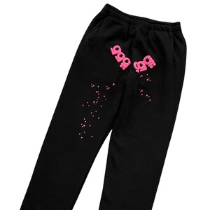 May include: Black sweatpants with a pink graphic design. The design includes the number "999" in a stylized font, repeated twice, and scattered pink star-shaped accents. The sweatpants have a drawstring waistband and pockets.