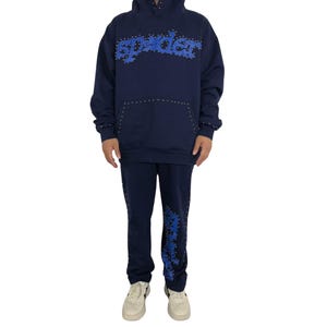 May include: Navy blue hoodie and sweatpants set. The hoodie features the word "spyder" in blue, studded with silver accents. The sweatpants have the same design on the left leg. The set includes a hood and a front pocket.