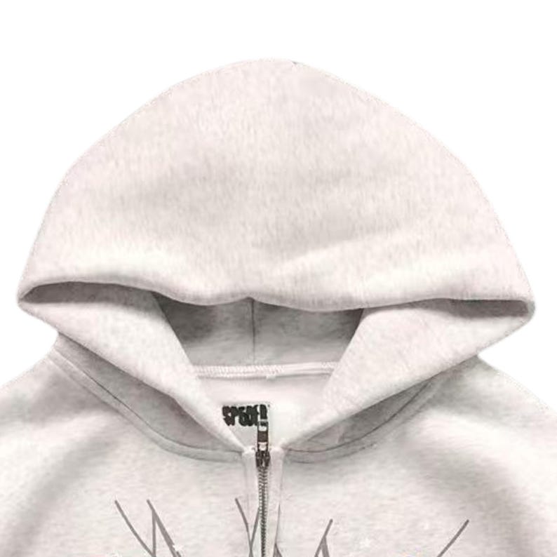 May include: Close-up of a light grey zip-up hoodie with a hood. The hoodie has a zip closure and a small logo near the neckline. The fabric appears to be a soft, fleecy material.