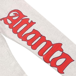 May include: A close-up shot of a light grey sweatpant leg featuring the word "Atlanta" in large, red, decorative lettering. The text is in a bold, stylized font, providing a strong contrast against the light grey fabric. The image focuses on the leg of the sweatpants.