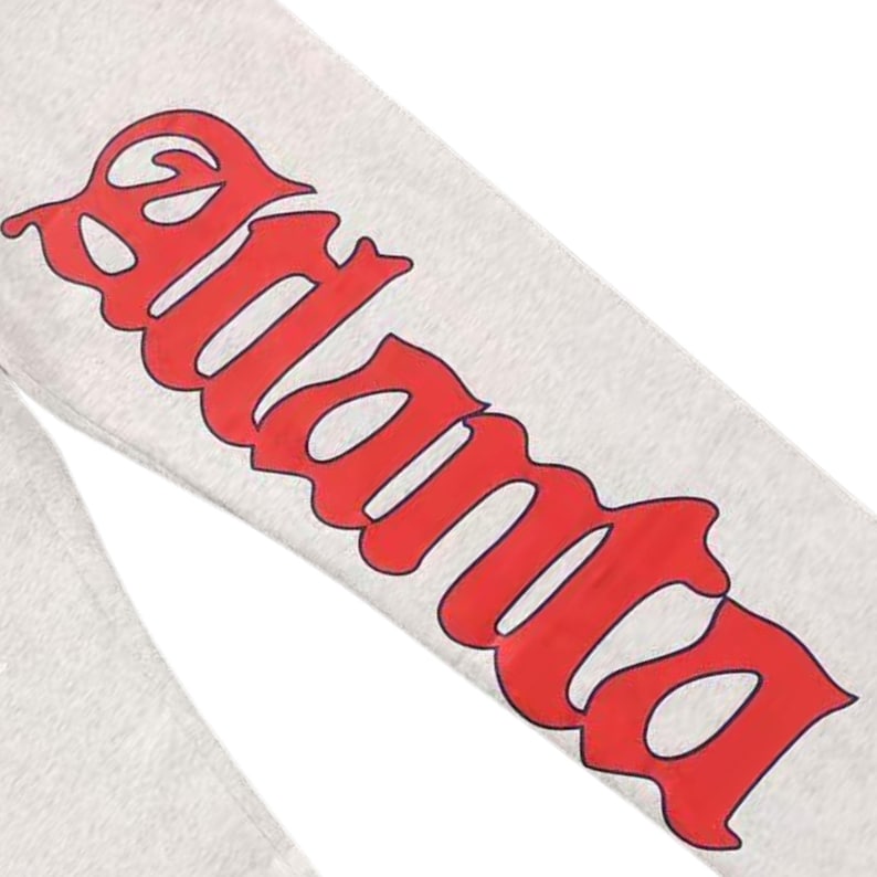 May include: A close-up shot of a light grey sweatpant leg featuring the word "Atlanta" in large, red, decorative lettering. The text is in a bold, stylized font, providing a strong contrast against the light grey fabric. The image focuses on the leg of the sweatpants.
