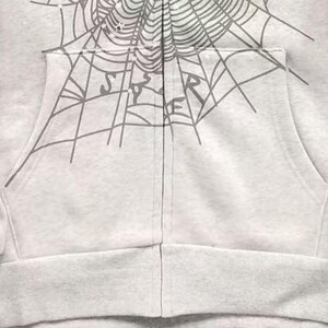 May include: A light grey zip-up hoodie with a grey spiderweb design across the top and the word "SPIDER" printed on the pocket. The hoodie has a front pocket and a ribbed hem.