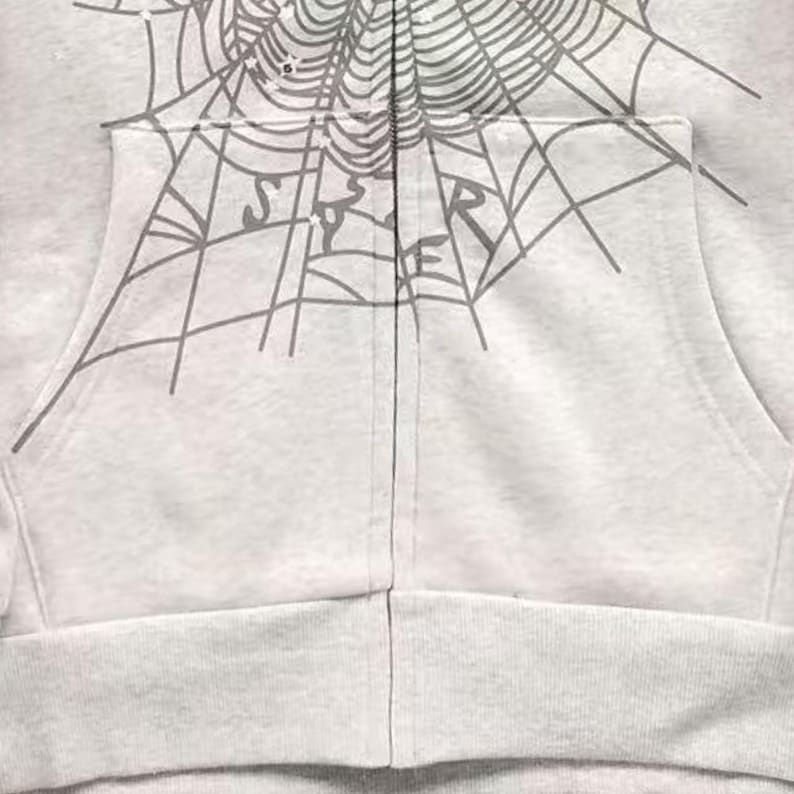 May include: A light grey zip-up hoodie with a grey spiderweb design across the top and the word "SPIDER" printed on the pocket. The hoodie has a front pocket and a ribbed hem.