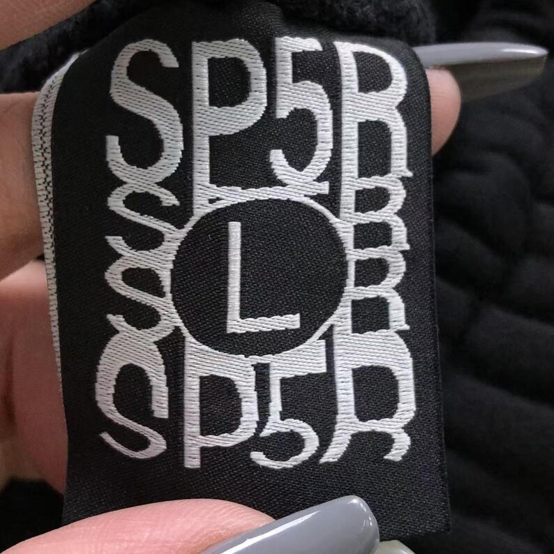 May include: Black fabric tag with white text. The tag features the letters "SP5R" and "SP5R" in a vertical arrangement, with a large "L" inside a circle in the center. The tag is held by a person.