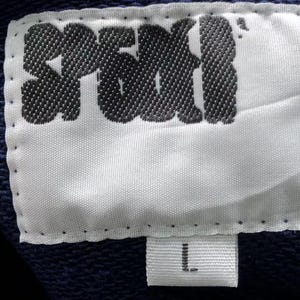 May include: Close-up of a white fabric label with the word "SPEKI" in black, stylized lettering. Below the main label is a smaller white tag with the letter "L" in black. The background is a dark blue fabric.