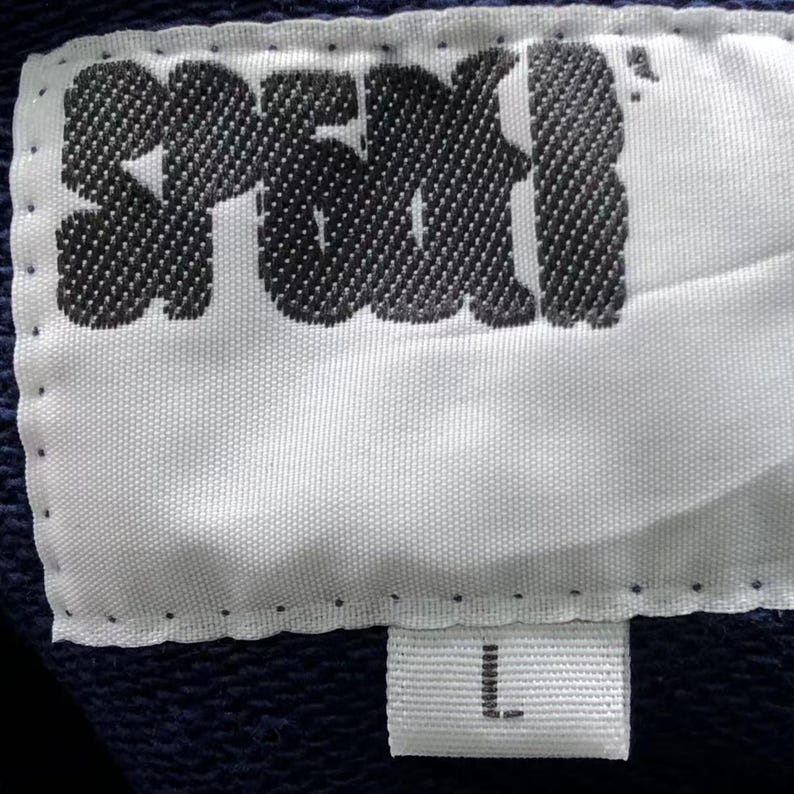 May include: Close-up of a white fabric label with the word "SPEKI" in black, stylized lettering. Below the main label is a smaller white tag with the letter "L" in black. The background is a dark blue fabric.