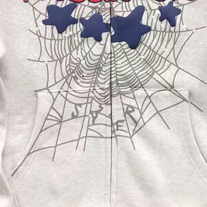 May include: A light grey zip-up hoodie with a spiderweb design and navy blue star accents. The word "STAR" is visible on the pocket. The hoodie has a front pocket and a zip closure.