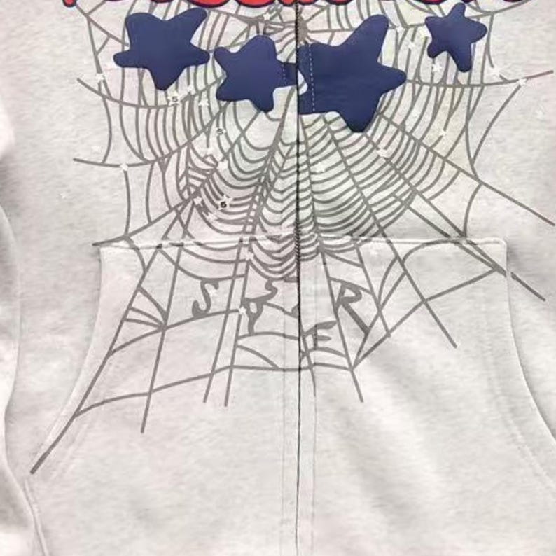 May include: A light grey zip-up hoodie with a spiderweb design and navy blue star accents. The word "STAR" is visible on the pocket. The hoodie has a front pocket and a zip closure.
