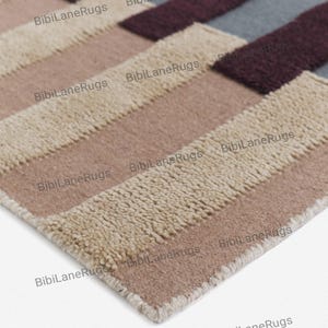 May include: Close-up of a rectangular rug with a geometric pattern. The rug features alternating stripes of beige, light brown, medium brown, burgundy, and grey. The rug has a soft, textured appearance and is suitable for home decor.