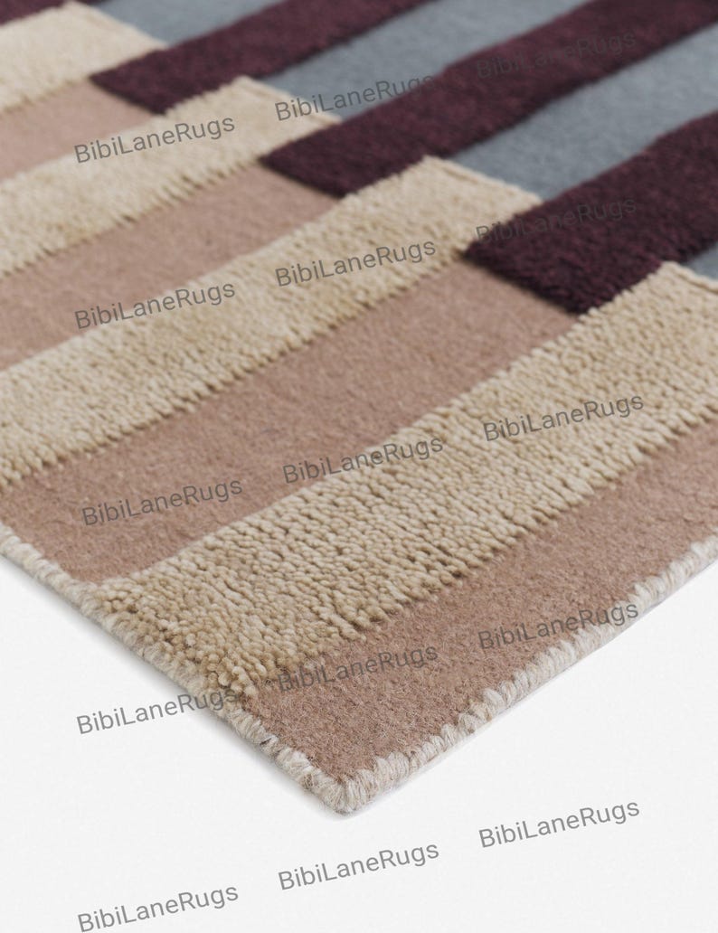 May include: Close-up of a rectangular rug with a geometric pattern. The rug features alternating stripes of beige, light brown, medium brown, burgundy, and grey. The rug has a soft, textured appearance and is suitable for home decor.