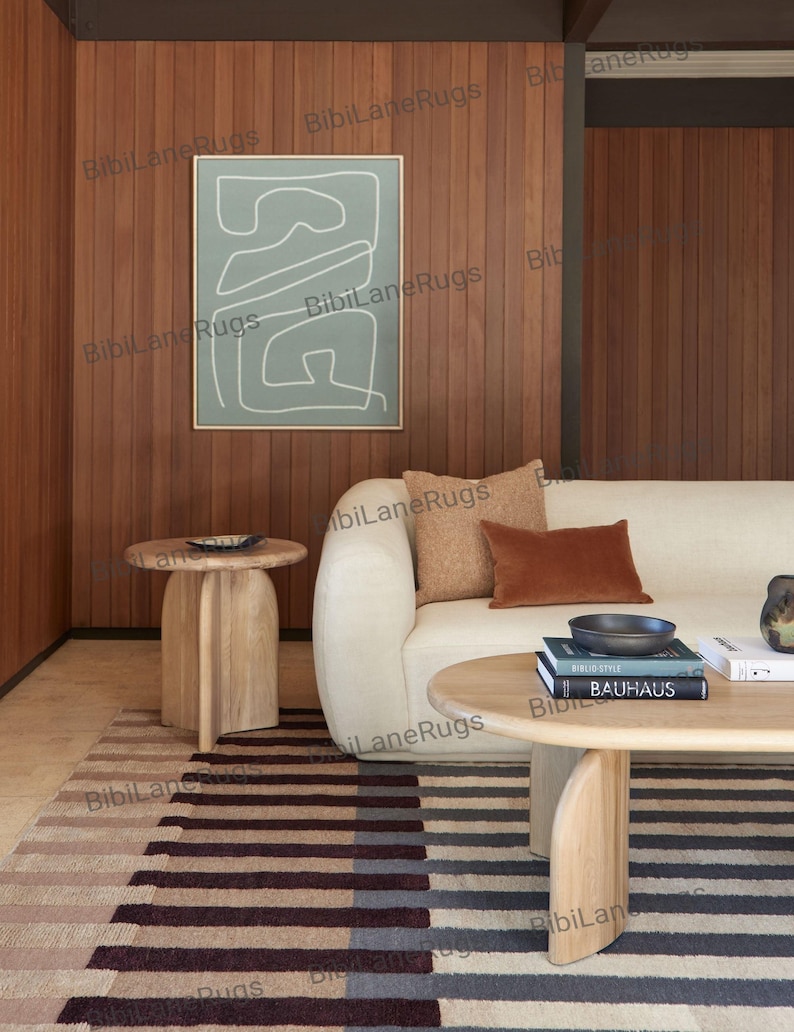 May include: A contemporary living room with a cream sofa and brown cushions. A light wood coffee table displays books and a bowl. A striped rug in brown, grey, and cream tones is on the floor. An abstract artwork hangs on the wood-panelled wall. The room has a minimalist design.