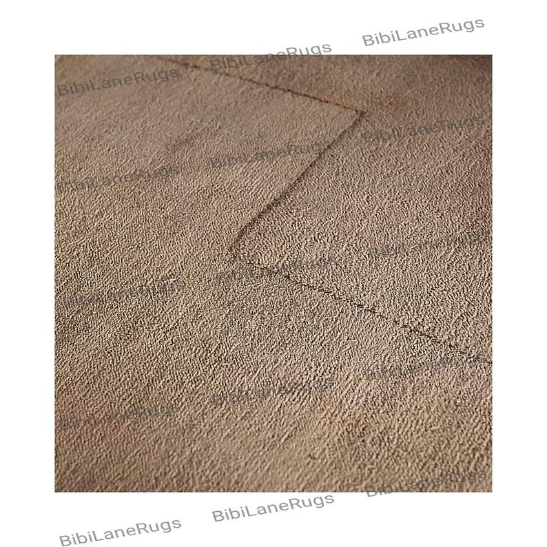May include: Close-up of a textured, beige rug with a geometric design. The rug has a soft, plush appearance and a subtle pattern created by the weave. The edges of the rug are slightly darker, creating a border effect. The rug is a neutral color.