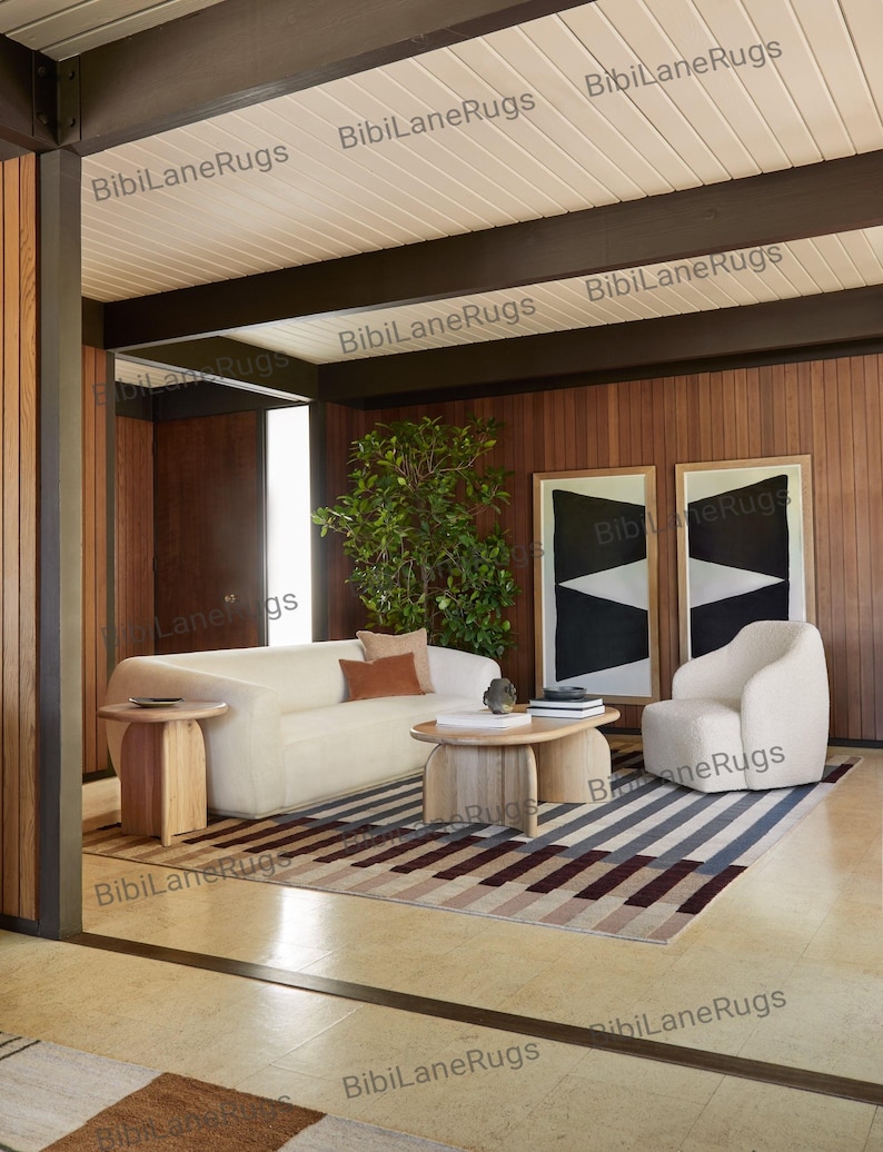 May include: A contemporary living room with a cream sofa, armchair, and a striped rug in grey, brown, and burgundy tones. Two framed abstract artworks adorn the wood-panelled wall. The room features wooden side tables and a coffee table. The room has a modern design.