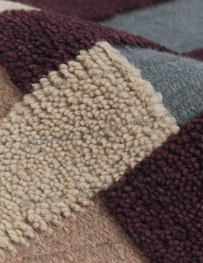 May include: Close-up of a section of a rug with a geometric pattern. The rug features a patchwork design with squares in shades of beige, grey, burgundy, and mauve. The texture is thick and plush, with visible loops and a soft, inviting appearance.