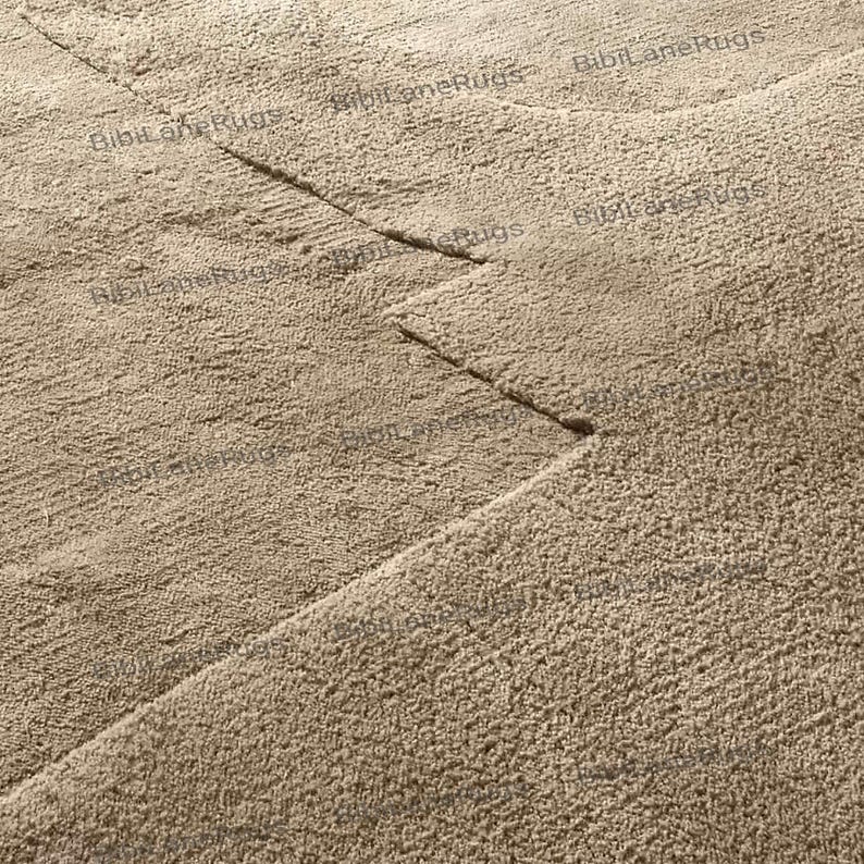 May include: Close-up of a beige rug with a textured surface. The rug has a geometric design with straight lines and angles. The rug is made of a soft, plush material, and the color is a neutral beige.