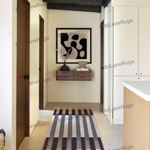 May include: A rectangular rug with a striped design in brown, beige, and blue tones. The rug is in a hallway with wooden doors and an abstract black and white artwork on the wall. The rug is likely 180 cm long.