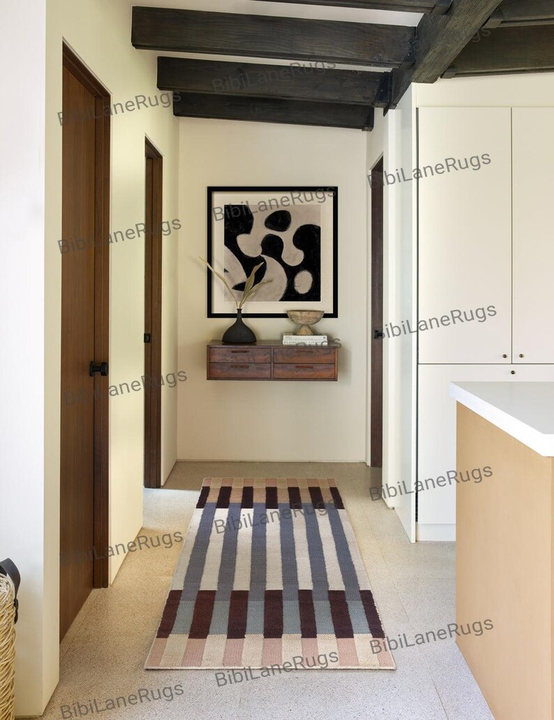 May include: A rectangular rug with a striped design in brown, beige, and blue tones. The rug is in a hallway with wooden doors and an abstract black and white artwork on the wall. The rug is likely 180 cm long.