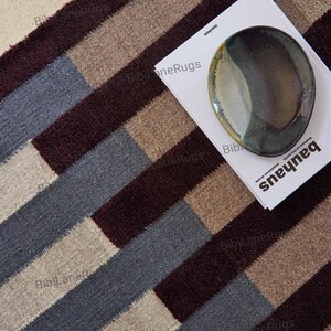 May include: A patterned rug featuring stripes in burgundy, grey, beige, and tan. A book with the word "bauhaus" and a decorative plate rest on the rug. The rug's design is geometric.