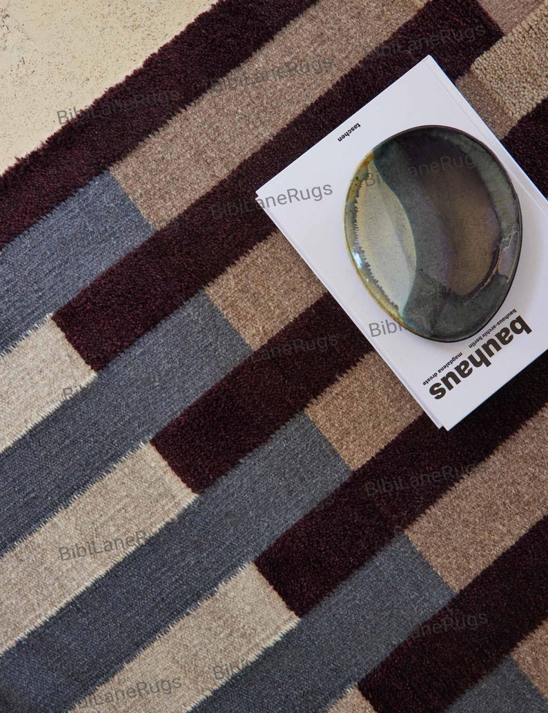 May include: A patterned rug featuring stripes in burgundy, grey, beige, and tan. A book with the word "bauhaus" and a decorative plate rest on the rug. The rug's design is geometric.