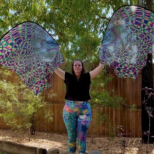 May include: A woman holding two large, colorful, patterned fabric fans. The fans have a psychedelic design with shades of purple, blue, green, and yellow. She is wearing a black top and leggings with a peacock feather design.