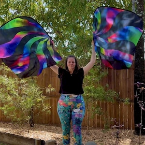 May include: Two large, colorful flags with swirling patterns of green, blue, purple, and pink are held aloft. The flags have a black border. The person in the image is wearing a black top and patterned leggings with peacock and floral designs.