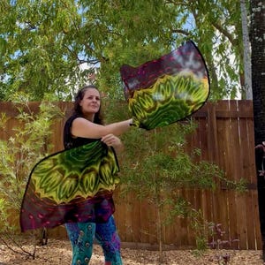 May include: A woman holding two colorful, patterned silk flags outdoors. The flags display a vibrant mix of green, yellow, and purple hues. The flags are in motion, creating a dynamic visual effect against a backdrop of greenery and a wooden fence.