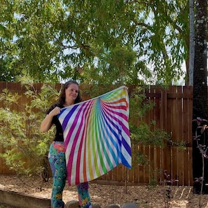 May include: A colorful, rainbow-striped scarf. The scarf features a vibrant design with radiating stripes of red, orange, yellow, green, blue, and purple. The scarf is held up against a backdrop of green foliage and a wooden fence.