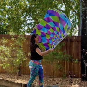 May include: A colorful, geometric-patterned flag in shades of purple, green, blue, and white is held aloft. The flag's design features a radial pattern. The person in the image is wearing a black top and patterned leggings.