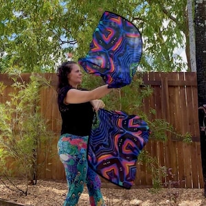 May include: A woman holding two colorful, patterned flags outdoors. The flags feature a vibrant design with blue, purple, orange, and black hues. She is wearing a black top and patterned leggings with a peacock design.