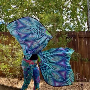 May include: A person wearing colorful peacock leggings and a black top, holding large, flowing, patterned wings. The wings feature a geometric design in shades of blue, purple, and green, creating a dynamic visual effect.