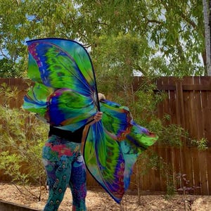 May include: Large, colourful butterfly wings in vibrant shades of blue, green, and yellow. The wings are held up against a background of green foliage and a wooden fence. The person is wearing peacock-themed leggings.