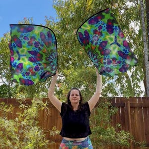 May include: Two colorful, patterned silk scarves are held up against a blue sky. The scarves feature a vibrant mix of green, blue, purple, and pink hues. The edges are outlined in black.