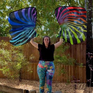 May include: Two large, colorful flags with a spiral design are held up by a person. One flag has a blue, purple, and black color scheme, while the other has a rainbow of colors. The flags appear to be made of a lightweight, flowing material.
