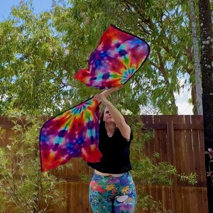 May include: Two colorful tie-dye flags with black borders are held up against a background of green foliage and a wooden fence. The flags display a vibrant mix of red, orange, yellow, green, blue, and purple hues. The flags are being waved in the air.