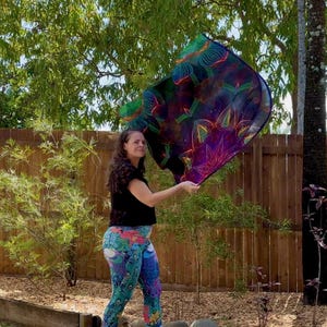 May include: A patterned scarf with a mandala design is held aloft. The scarf displays shades of purple, green, and blue. The person is wearing a black top and peacock-print leggings.