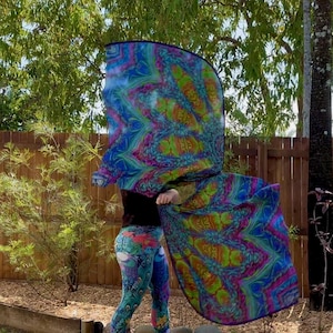 May include: A vibrant, patterned scarf in shades of blue, green, and pink is held up in the air. The scarf's design is mirrored, creating a symmetrical, psychedelic effect. The person is wearing patterned leggings and a black top.